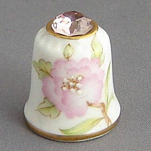 May include: A white porcelain thimble with a pink flower design and a clear rhinestone on top.