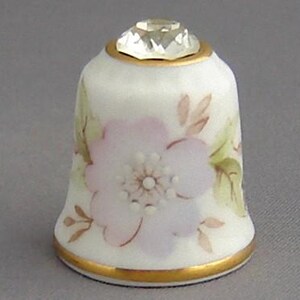 May include: A white porcelain thimble with a gold trim and a single pink flower with a white center. The thimble has a clear rhinestone on top.
