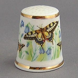 May include: A white porcelain thimble with a gold trim and a painted image of a yellow and black butterfly with blue flowers.