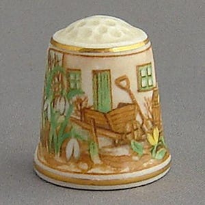 May include: A white porcelain thimble with a gold trim and a painted scene of a wheelbarrow with flowers and a small house.
