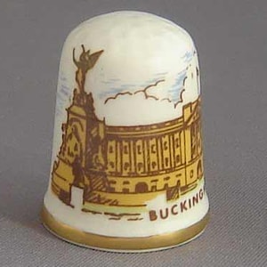 Puede incluir: Un dedal de cerámica blanca con un borde dorado. El dedal presenta una ilustración marrón y amarilla del Palacio de Buckingham con el texto "BUCKING" visible en la parte inferior.