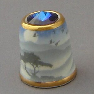 May include: A blue and white porcelain thimble with a gold rim and a blue crystal on top. The thimble features a landscape scene with a moon, trees, and birds flying in the sky.