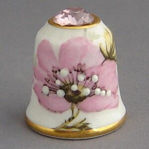 May include: A white porcelain thimble with a pink flower design and a pink rhinestone on top. The thimble has a gold trim around the edge.