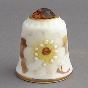May include: White porcelain thimble with a gold trim and a brown and white floral design. The thimble has a clear amber gem on top.