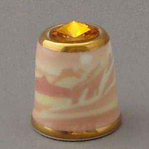 May include: A pink and white ceramic thimble with a gold trim and a yellow rhinestone on top.