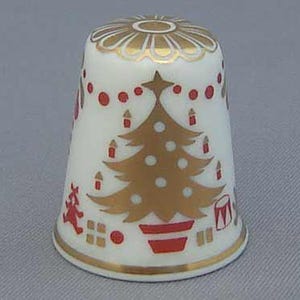 Puede incluir: Un dedal de porcelana blanca con un diseño de árbol de Navidad dorado y rojo. El dedal presenta un borde dorado y un patrón floral dorado en la parte superior. Puntos rojos y elementos festivos completan el tema navideño.