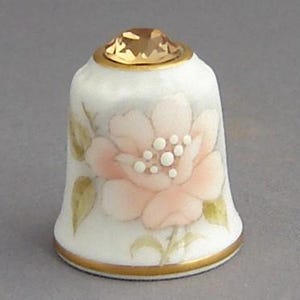 May include: White porcelain thimble with a pink flower design and a gold trim. The thimble has a clear crystal on top.