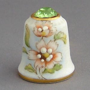 May include: A white porcelain thimble with a floral design and a green gemstone on top. The thimble is decorated with pink and white flowers and green leaves.