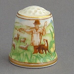 May include: A porcelain thimble with a hand-painted scene of a shepherd with a flock of sheep and a dog in a green field. The thimble has a gold trim around the top and bottom.