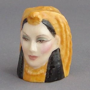 May include: A ceramic thimble featuring a woman's face with a yellow and black headdress. The woman has green eyes and a light pink blush on her cheeks.