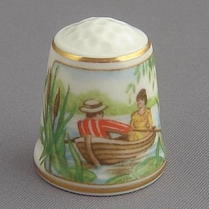May include: A porcelain thimble with a painted scene of a couple in a rowboat on a lake. The thimble is decorated with green reeds and lily pads.