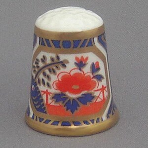 Puede incluir: Un dedal de porcelana blanca con un diseño floral azul y dorado. El dedal presenta una flor roja con hojas azules y un borde dorado.