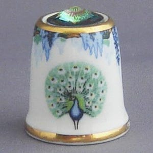 May include: A white porcelain thimble with a gold trim and a green and blue peacock design. The thimble has a clear iridescent jewel on top.