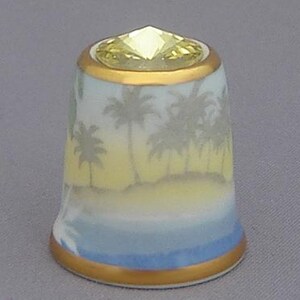 May include: A white porcelain thimble with a yellow and blue tropical scene. The thimble has a gold rim and a clear crystal top.