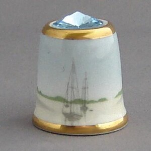 May include: A white porcelain thimble with a gold trim and a blue crystal top. The thimble features a painted scene of a sailboat on the water.