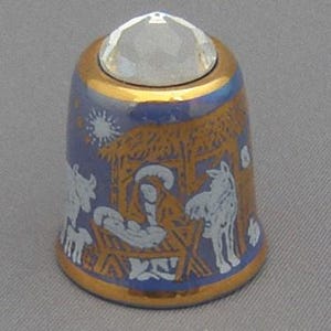 May include: A blue and gold ceramic thimble with a crystal top. The thimble features a white depiction of the nativity scene with a star above the stable.