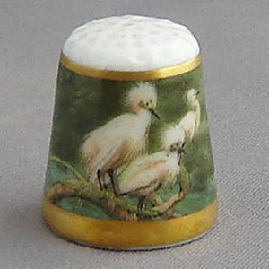 May include: White porcelain thimble with a gold rim and a green and gold design featuring three white birds with long legs and beaks.