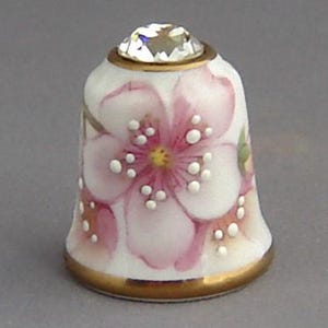 May include: A white porcelain thimble with a pink floral design and a clear crystal top.