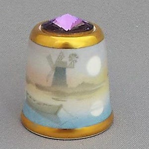 May include: A porcelain thimble with a gold rim and a purple gemstone on top. The thimble features a painted scene of a windmill and a boat on the water.