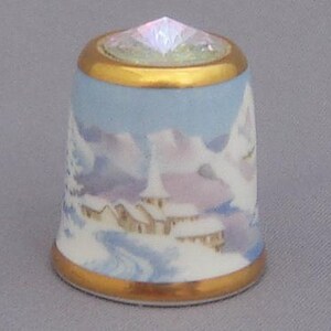 May include: A white porcelain thimble with a gold trim and a winter scene. The scene features a snowy landscape with a church and trees. The thimble has a clear iridescent crystal on top.