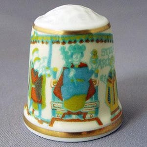 May include: A white porcelain thimble with a gold trim and a colorful scene of people and a building. The text "St. Andrews" is visible on the thimble.
