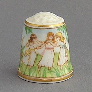 May include: A porcelain thimble with a hand-painted design of three girls in a field of flowers. The girls are wearing white dresses and have blonde hair. The thimble is decorated with gold trim.