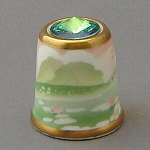 May include: A white porcelain thimble with a green and pink landscape scene. The thimble has a gold rim and a green gemstone on top.