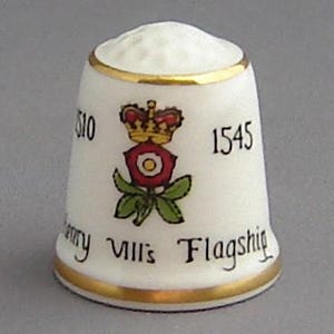 May include: White porcelain thimble with a gold trim and a red and green floral design. The thimble has the number 1545 and the text "Henry VIII's Flagship" printed on it.