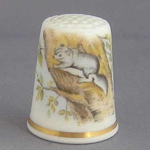 May include: A white ceramic thimble with a gold trim. The thimble features a painted image of a small, gray animal sitting on a tree branch.
