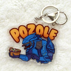 Puede incluir: Llavero de acrílico transparente con un personaje de dibujos animados azul sosteniendo un cuenco de comida, con la palabra "POZOLE" en letras naranjas. El llavero tiene un llavero y un cierre de metal plateado.