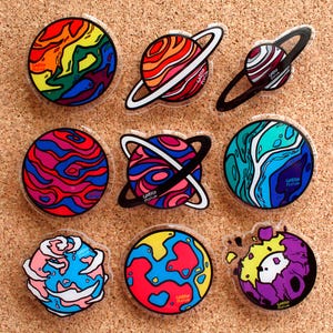 May include: Nine colourful planet-shaped acrylic pins with unique designs and the text "Gaby Plutan". The pins feature vibrant colours like red, blue, yellow, and purple, with some having rings around them. They are arranged on a corkboard.