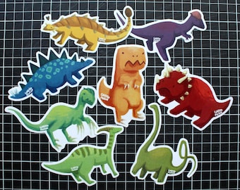 May include: A collection of colorful dinosaur stickers in various shapes and sizes. The stickers feature illustrations of different dinosaurs in shades of yellow, blue, orange, green, and red. The stickers are on a black grid background.