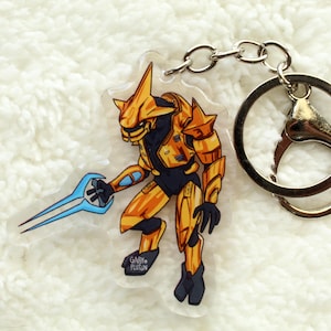 Puede incluir: Un llavero Halo Elite dorado y azul con un llavero plateado. El Elite sostiene una espada de energía azul.