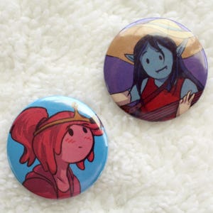 May include: Two round button badges featuring characters from the animated series Adventure Time. The first badge features a blue-skinned character with long black hair wearing a red shirt and a straw hat. The second badge features a pink-skinned character with long red hair wearing a pink shirt and a gold crown.