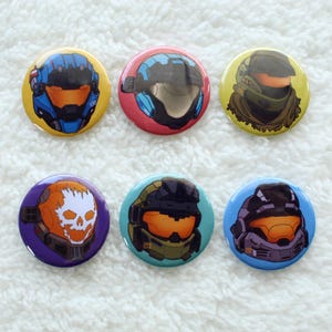 Puede incluir: Seis botones redondos con fondos de colores diferentes e imágenes de personajes con cascos. Los personajes están representados en un estilo de dibujos animados. Los botones tienen un diámetro de aproximadamente 2,5 cm.