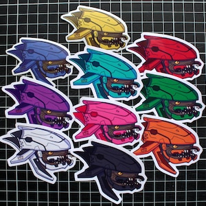 Może przedstawiać: Dziesięć kolorowych naklejek winylowych z kreskówkową ilustracją hełmu o groźnym wyrazie. Hełmy są w różnych kolorach, w tym żółtym, czerwonym, niebieskim, fioletowym, różowym, zielonym, pomarańczowym, białym i czarnym.