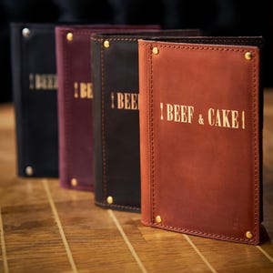 May include: A row of leather menu covers in various colors: black, burgundy, dark brown, and reddish-brown. Each cover is embossed with the words "BEEF & CAKE" in gold lettering. The covers are secured with brass-colored rivets.