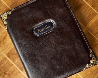 May include: A dark brown leather journal with the name "Emilia" embossed on a navy blue patch. The journal has decorative gold-coloured corner accents and is set on a wooden surface.
