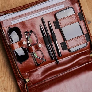 May include: A brown leather portfolio case with a zip fastening. The case is open and contains a smartphone, glasses, and pens. The case has the text "Robert Smith" embossed on the front.