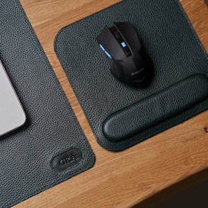 Könnte beinhalten: Ein dunkelgrünes Leder-Schreibtischpad mit passendem Mauspad und Handgelenkauflage. Eine schwarze kabellose Maus mit blauen Akzenten liegt auf dem Mauspads. Ein Laptop ist teilweise auf dem Schreibtischpad zu sehen. Der Schreibtisch ist aus Holz.