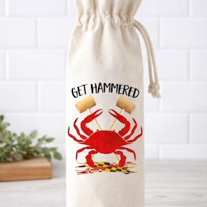 Op de afbeelding: Een beige wijnfleszak met een trekkoordsluiting. De tas heeft de tekst "GET HAMMERED" boven een rode krabafbeelding met twee houten hamers. De krab staat boven een Maryland-vlagontwerp.