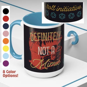 May include: A white ceramic mug with a blue handle. The mug has a black background with a red and gold illustration of a treasure chest with teeth and the text "Definitely Not a Mimic". The mug has a black band around the top with the text "roll initiative" and five blue dice.