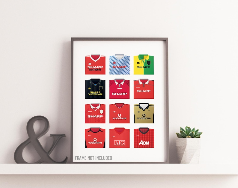 May include: A framed print featuring a collection of twelve soccer jerseys in various colors, including red, blue, and yellow. Each jersey displays sponsor logos such as "SHARP" and "Vodafone". The phrase "FRAME NOT INCLUDED" is printed at the bottom.