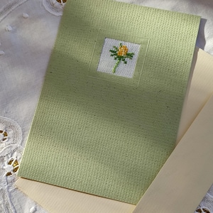 Könnte beinhalten: Eine handgefertigte Grußkarte mit hellgrün strukturierter Hülle. Die Karte zeigt ein kleines, quadratisches Kreuzstichmuster einer gelben Blume mit grünen Blättern. Ein cremefarbener Umschlag ist enthalten.