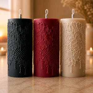 Peut inclure: Trois bougies cylindriques en noir, rouge foncé et crème. Chaque bougie présente un motif floral en relief rappelant de la dentelle, enveloppant l'extérieur, avec des détails de dentelle suspendus en bas et une petite boucle métallique argentée au sommet.