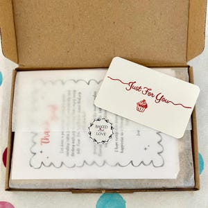May include: A brown cardboard box containing a white card with the red text Just For You and a cupcake illustration. A small circular sticker reads Baked with LOVE. Beneath these, a translucent paper sheet features a scalloped edge and text.