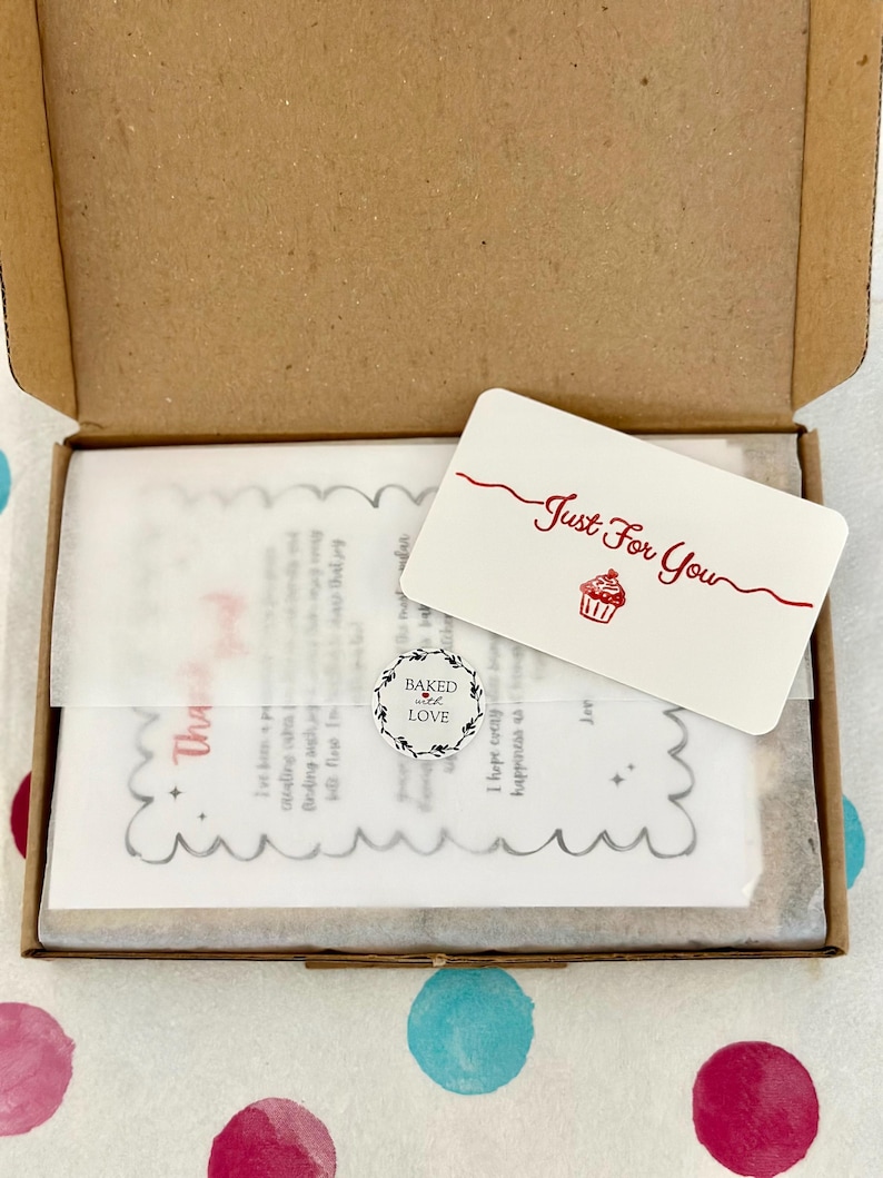 May include: A brown cardboard box containing a white card with the red text Just For You and a cupcake illustration. A small circular sticker reads Baked with LOVE. Beneath these, a translucent paper sheet features a scalloped edge and text.