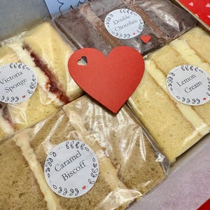 May include: A gift box filled with four different types of cake slices. The cakes are wrapped in clear plastic and labelled with their flavours: Victoria Sponge, Double Chocolate, Caramel Biscoff, and Lemon Cream. A red heart is placed on top of the box.