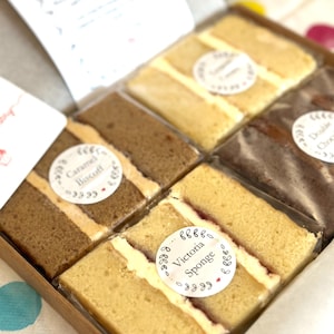 May include: A cardboard box containing four rectangular slices of cake, each individually wrapped in clear plastic. The flavors include Victoria Sponge, Caramel Biscoff, and Double Chocolate, each labeled with a round white sticker.