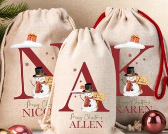 Puede incluir: Tres bolsas de regalo navideñas beige con cordones. Cada bolsa presenta una gran letra burdeos, una ilustración de muñeco de nieve y las palabras "Feliz Navidad" con un nombre. Las bolsas tienen cordones rojos o beige.